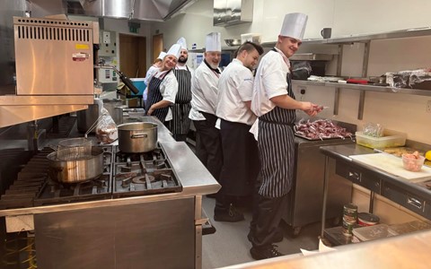 A group of chefs in white uniforms and hats work in a professional kitchen with stainless steel appliances. One chef smiles at the camera while others focus on preparing food. Ingredients and utensils are spread across the countertops