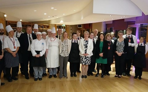A group of people standing in a room with wooden floors and a curved ceiling. Some are dressed in chef uniforms with white hats, while others wear formal or business casual attire. The background features shelves and various lighting fixtures