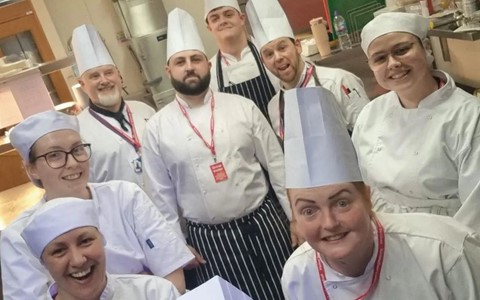 A group of chefs wearing white uniforms and tall white hats standing together in a commercial kitchen. Some chefs are giving thumbs up. The kitchen features stainless steel appliances, shelves stocked with various items, and is brightly lit by overhead lighting