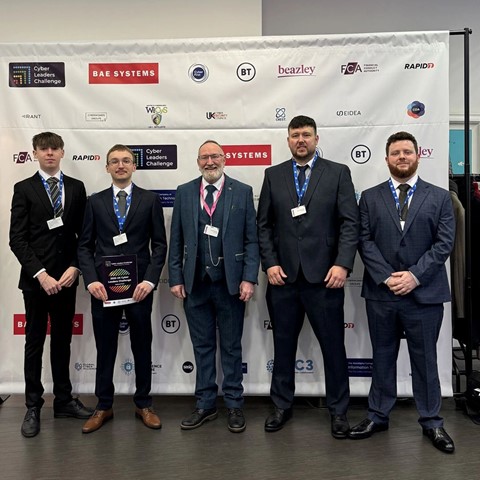 Five individuals in formal attire stand in front of a backdrop displaying logos such as 'BAE Systems,' 'beazley,' 'FCA,' and 'BT.' They are wearing name badges, and one person is holding a plaque or award. The backdrop indicates the event is the 'Cyber Leaders Challenge.