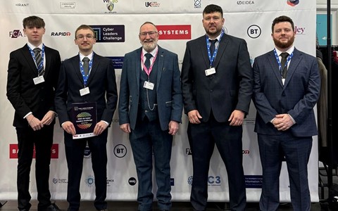 Five individuals in formal attire stand in front of a backdrop displaying logos such as 'BAE Systems,' 'beazley,' 'FCA,' and 'BT.' They are wearing name badges, and one person is holding a plaque or award. The backdrop indicates the event is the 'Cyber Leaders Challenge.