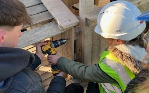 A group of people working on a wooden structure outdoors in daylight. Some are wearing high-visibility vests, and one person is wearing a white hard hat. One individual is using a yellow and black power drill on the structure.