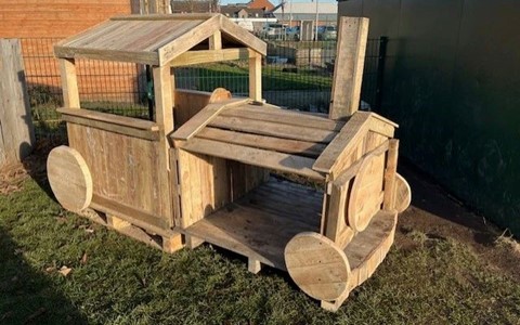 A wooden play structure shaped like a train engine, featuring a roofed section at the back, open sides, and large circular wooden wheels. It is placed outdoors on grass, with a fence and buildings visible in the background.