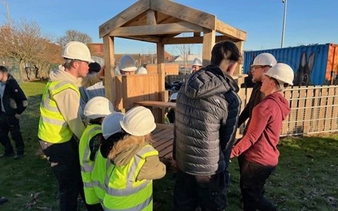A group of children and adults wearing hard hats and high-visibility vests work together to carry a wooden structure resembling a small gazebo or playhouse. The scene is set outdoors on grass, with trees and buildings in the background