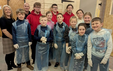 A group of children and adults standing indoors in front of a wooden structure. The children are wearing blue plastic aprons and white gloves, with some holding paintbrushes. A green paint bucket is on the floor in front of them. The adults, dressed in red hoodies, stand behind the children