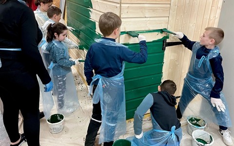 Children and adults painting a wooden structure with green paint. The children are wearing blue aprons and gloves, using paintbrushes to apply the paint. Several green paint buckets are on the floor, and the structure—possibly a shed—is partially painted