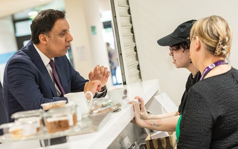 Three people interacting at a counter in a café or food service area. One person in a suit and tie leans on the counter, speaking with two others—one wearing a black cap with visible arm tattoos, and the other with blonde hair tied back, glasses, and a lanyard. The counter holds food items in plastic containers, cups, and small objects. The background includes windows and doors in an indoor setting
