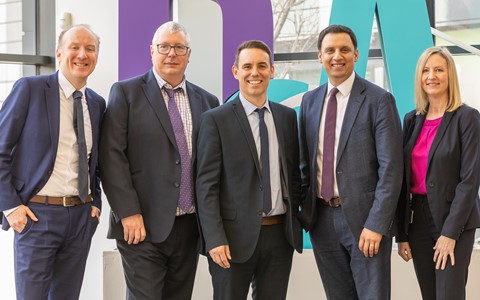 Five individuals in business attire stand in a row indoors, in front of large, teal and purple D&A letters.