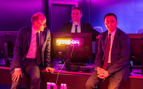 Three individuals in suits, are positioned around a desk with computer monitors. The room is lit with purple and pink lighting, and one monitor displays a neon sign that reads 'game on'.