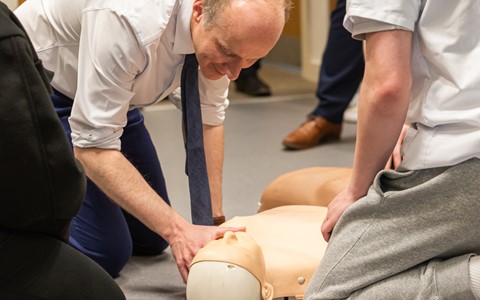 A group of people gathered around a CPR training mannequin on the floor. One person in a white shirt and tie is demonstrating chest compressions, while others observe closely. One individual kneels beside the instructor. The setting is indoors, with additional people standing and watching in the background.