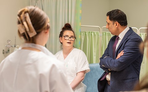 A group of four people in a medical setting inspired classroom. Three individuals are wearing white coats, and one is in a suit. The background features green curtains and medical equipment.