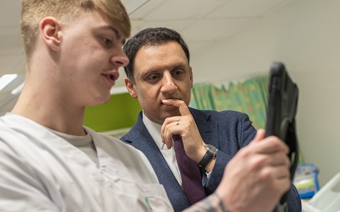 Two individuals in a medical setting. One is wearing a white medical uniform and holding a device, possibly a tablet or medical equipment. The other is in formal attire with a suit and tie, appearing to observe or discuss something