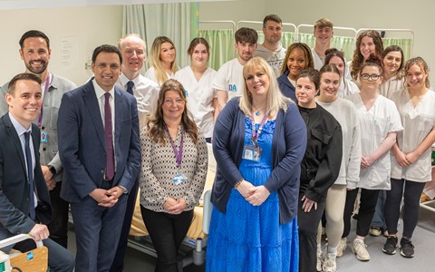 A group of men and women standing in a room resembling a medical or healthcare setting. Some are in professional attire like suits, while others wear white uniforms typical of healthcare workers. The background features hospital beds and privacy curtains.