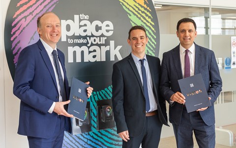 Three individuals in business attire stand in front of a large circular sign featuring colorful fingerprint patterns and the text 'the place to make your mark.' Each person holds a dark blue folder with a logo and text. The background shows an office setting with glass walls and doors