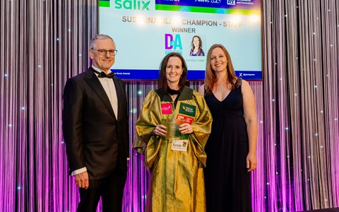 Three people standing in front of a backdrop with vertical silver strings. The person in the centre, wearing a green robe, holds an award. The person on the left is in a black tuxedo, and the person on the right is in a dark blue dress. Behind them, a screen displays the text: '20th Anniversary Green Gown Awards 2024 UK Research and Innovation – SUSTAINABILITY CHAMPION - STAFF WINNER – Christine Calder.'"