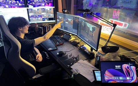 A person seated in a gaming chair at a desk with multiple monitors showing video feeds and editing software. They are pointing at one of the screens. The desk holds energy drink cans, a keyboard, mouse, smartphone, and other electronic gear.