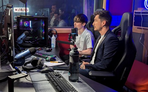Two people seated in front of a computer setup with multiple monitors, microphones, and a gaming PC. The room features a professional studio setup with an illuminated 'ON AIR' sign in the background.
