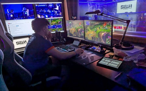 A person seated at a desk with multiple monitors displaying a video game and other content. They are using a keyboard and mouse, with a microphone, tablet, and various papers also on the desk. In the background, a room with blue lighting and a large screen showing the text 'SELI' is visible through a window.