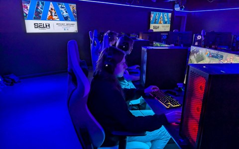 A dimly lit gaming room with blue ambient lighting. Several people are seated in ergonomic gaming chairs, wearing headsets, and playing games on desktop computers. The monitors display various game scenes.