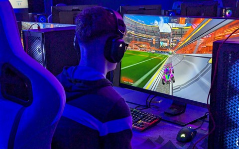 A person wearing headphones sits in an ergonomic gaming chair, playing Rocket League on a computer monitor. The game shows a car on a soccer field. The setting is a gaming or esports environment with multiple computer setups and blue ambient lighting