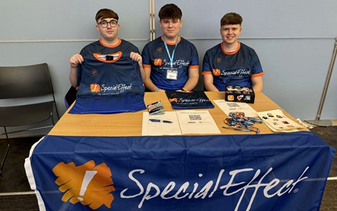 Three individuals sit behind a table covered with a blue 'SpecialEffect' tablecloth. They are wearing matching blue shirts with orange accents, also branded with the 'SpecialEffect' logo. The person on the left is holding up another shirt. The table displays flyers, a QR code, lanyards, and a box of promotional materials