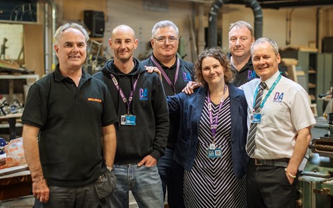 A group of six people standing together in a workshop or industrial setting. They are wearing various types of clothing, including polo shirts, hoodies, and a dress with a blazer. Each person has an identification badge on a lanyard around their neck. The background shows workbenches, tools, and machinery typically found in a workshop environment