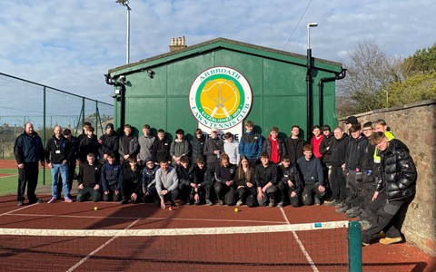 Group photo of around 40 people on a red clay tennis court in front of a green building labeled 'Arbroath Lawn Tennis Club.' Some individuals are standing while others are crouching, all in casual attire. A tennis net is visible in the foreground under a partly cloudy sky