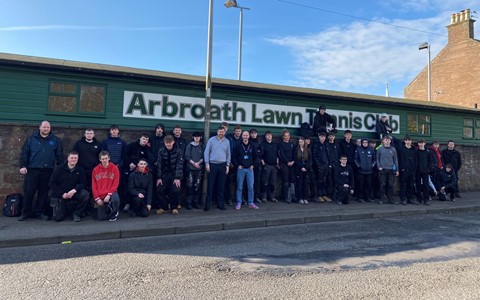 Group photo of people standing and sitting in front of a green building with a sloped roof and a sign that reads 'Arbroath Lawn Tennis Club.' Street lamps and a clear blue sky are visible in the background