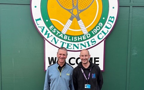 Two individuals stand in front of a large sign for the Arbroath Lawn Tennis Club, which features two crossed tennis rackets and the text 'Arbroath Lawn Tennis Club Established 1909' in green and yellow. A smaller white sign below reads 'Welcome.' One person wears a light blue jacket, black pants, and white shoes; the other wears a black hoodie, light blue jeans, and pink shoes. They are on a red surface with green walls behind them.
