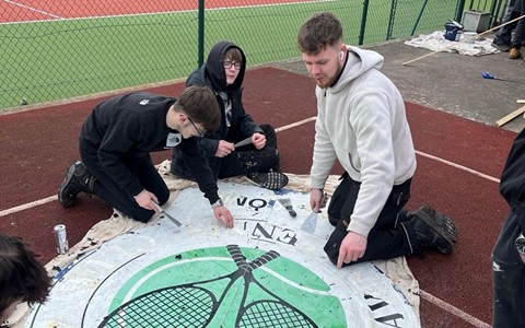 A group of people painting a large circular sign on the ground near a fenced tennis court with red and green surfaces. The sign features two crossed black tennis rackets over a green circle, with the text 'ARBROATH LAWN TENNIS CLUB' written around the edge.