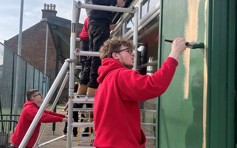 Four individuals painting the exterior of a building. Three are on a scaffold and one is on the ground. All are wearing red hoodies and black pants. One person is using a brush to paint a green section of the wall. A tall streetlight and part of another building are visible in the background