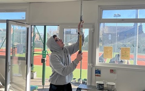 A person in a gray hoodie and black pants painting a ceiling with a roller on an extension pole. The room has large windows showing a tennis court outside. Paint cans, a step ladder, and other supplies are on the floor, which is covered with drop cloths