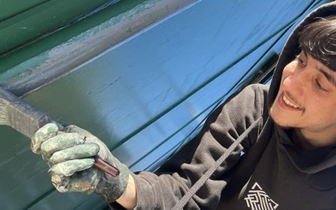 A person in a black hoodie and gloves painting a green metal surface with a paintbrush. Their face is partially obscured. Sunlight and shadows are visible in the outdoor background