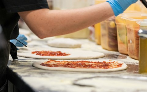 A person spreads pizza sauce on a pizza base. The base is on a floured surface