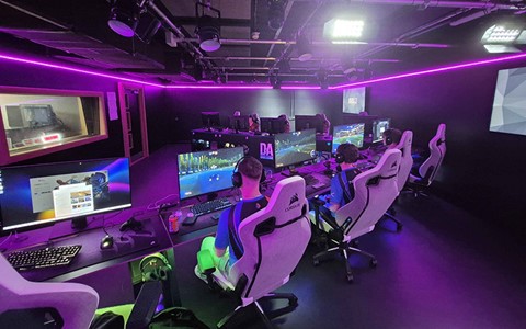 A gaming room with multiple computer stations and purple LED lighting. Several individuals are seated in gaming chairs, wearing headsets and focused on their monitors.