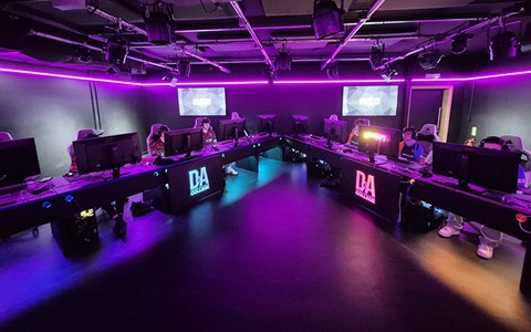 A wide-angle view of a gaming room with rows of computer stations facing each other, illuminated by purple LED lights.