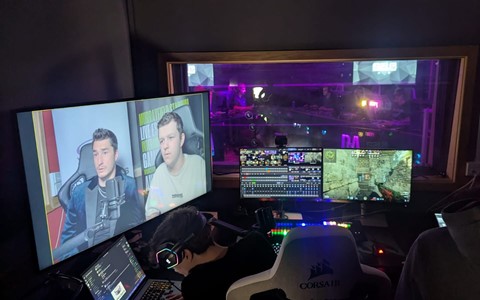 A control room setup with two large monitors showing a live broadcast of two speakers. A person wearing headphones is seated at a desk with a laptop, surrounded by smaller screens and control panels.
