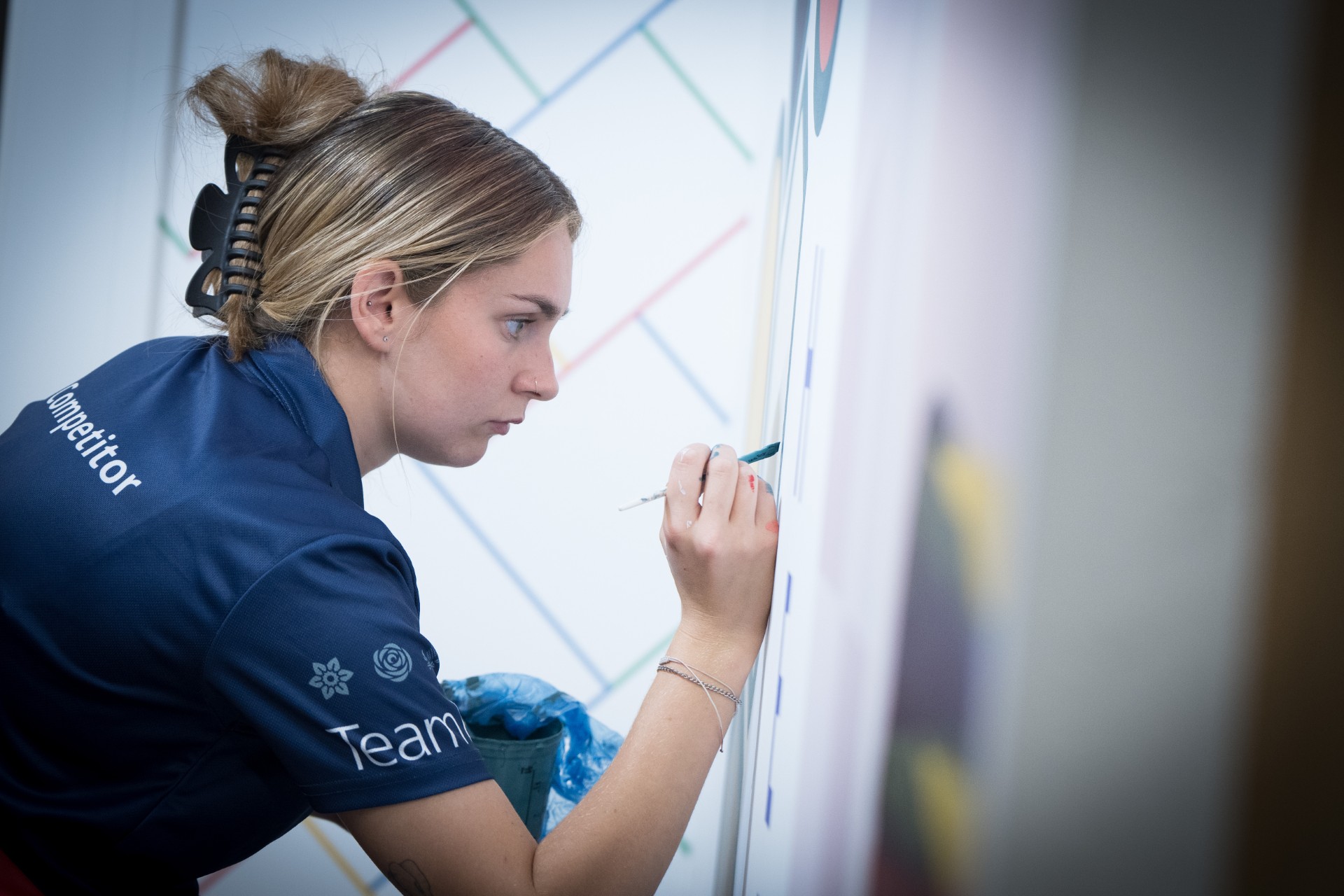 A person in a blue team shirt is painting fine details on a white wall with a small brush. The wall features a colorful diagonal line pattern, and the person is holding a paint container in the other hand.