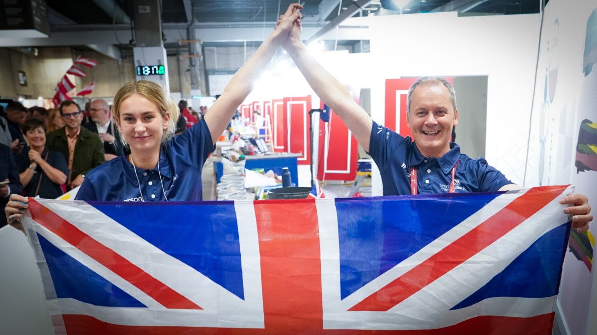 Two individuals wearing matching navy shirts hold up a large Union Jack flag in front of them, with their hands raised and clasped together in celebration. The background shows a busy indoor event space with red panels and other people.)