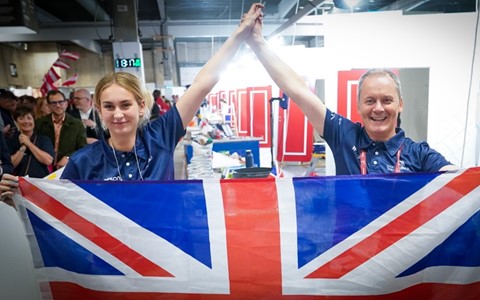 Two individuals wearing matching navy shirts hold up a large Union Jack flag in front of them, with their hands raised and clasped together in celebration. The background shows a busy indoor event space with red panels and other people.
