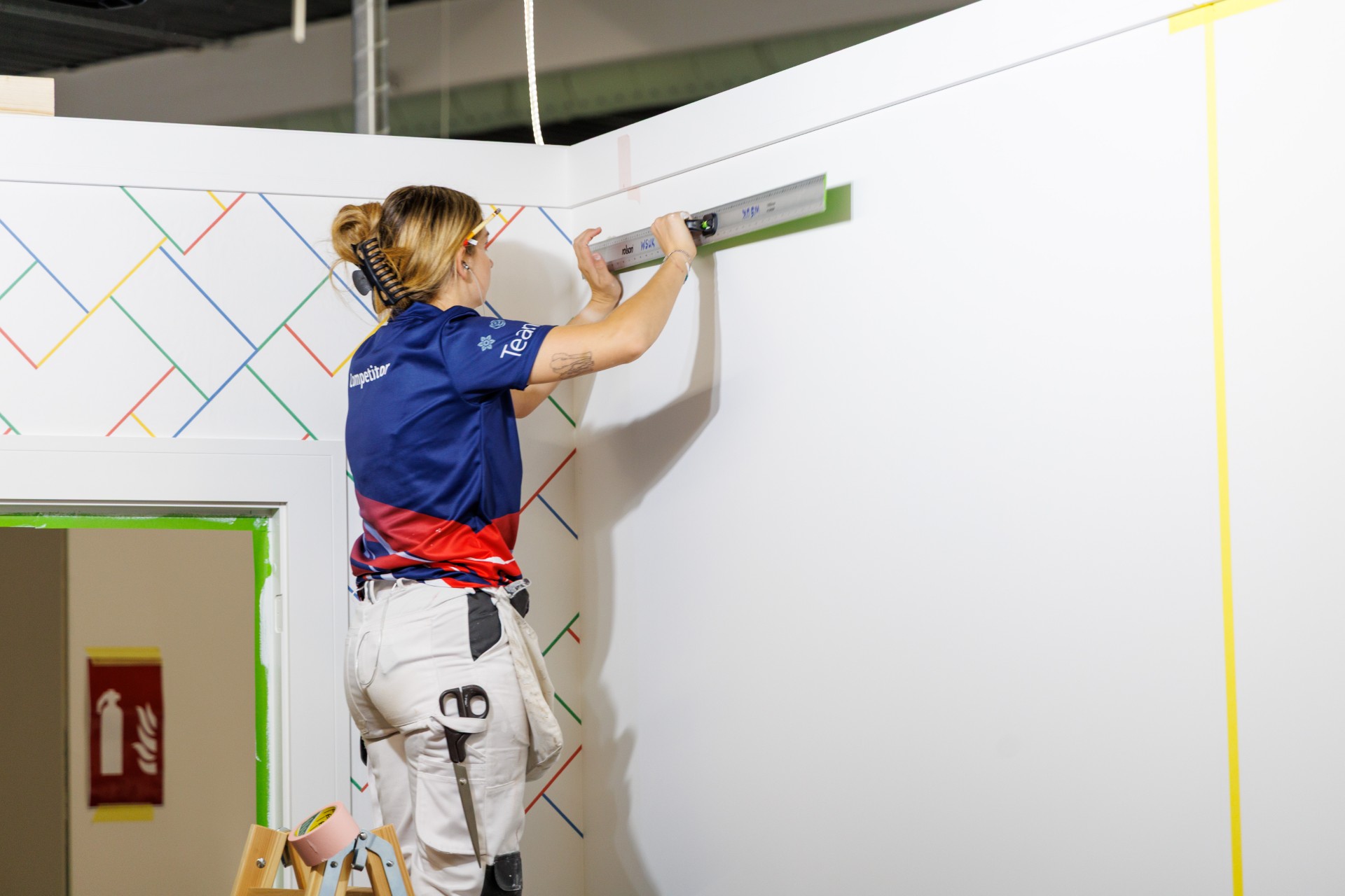 A person in a blue and red team shirt stands on a stepladder, using a spirit level to check alignment on a white wall decorated with a colorful diagonal line pattern. A doorway with green tape around the frame is visible below.