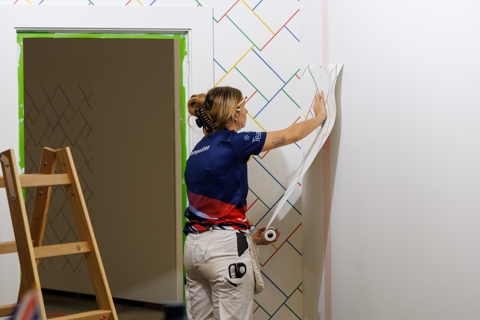 A person in a blue and red team shirt is applying wallpaper to a white wall with a colourful diagonal line pattern. A doorway with green tape and a wooden stepladder are visible nearby.