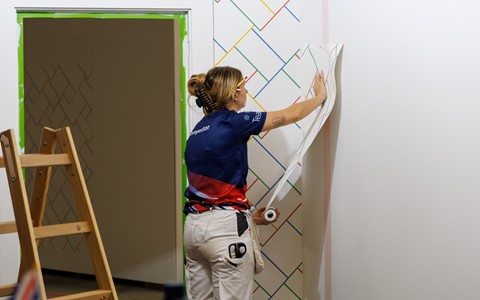 A person in a blue and red team shirt is applying wallpaper to a white wall with a colourful diagonal line pattern. A doorway with green tape and a wooden stepladder are visible nearby.