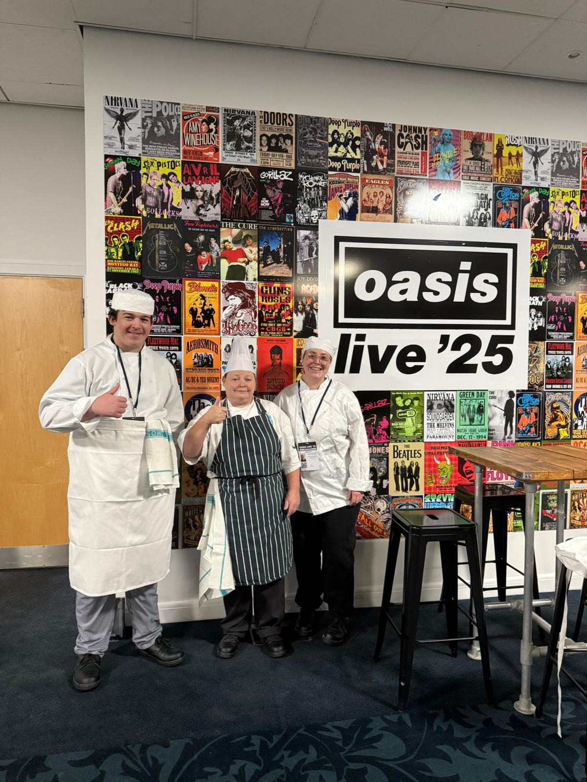 Three individuals dressed in chef uniforms are standing in front of a colorful wall covered with music posters. A large sign in the centre reads “oasis live ’25.” There is a tall wooden table with black bar stools to the right.