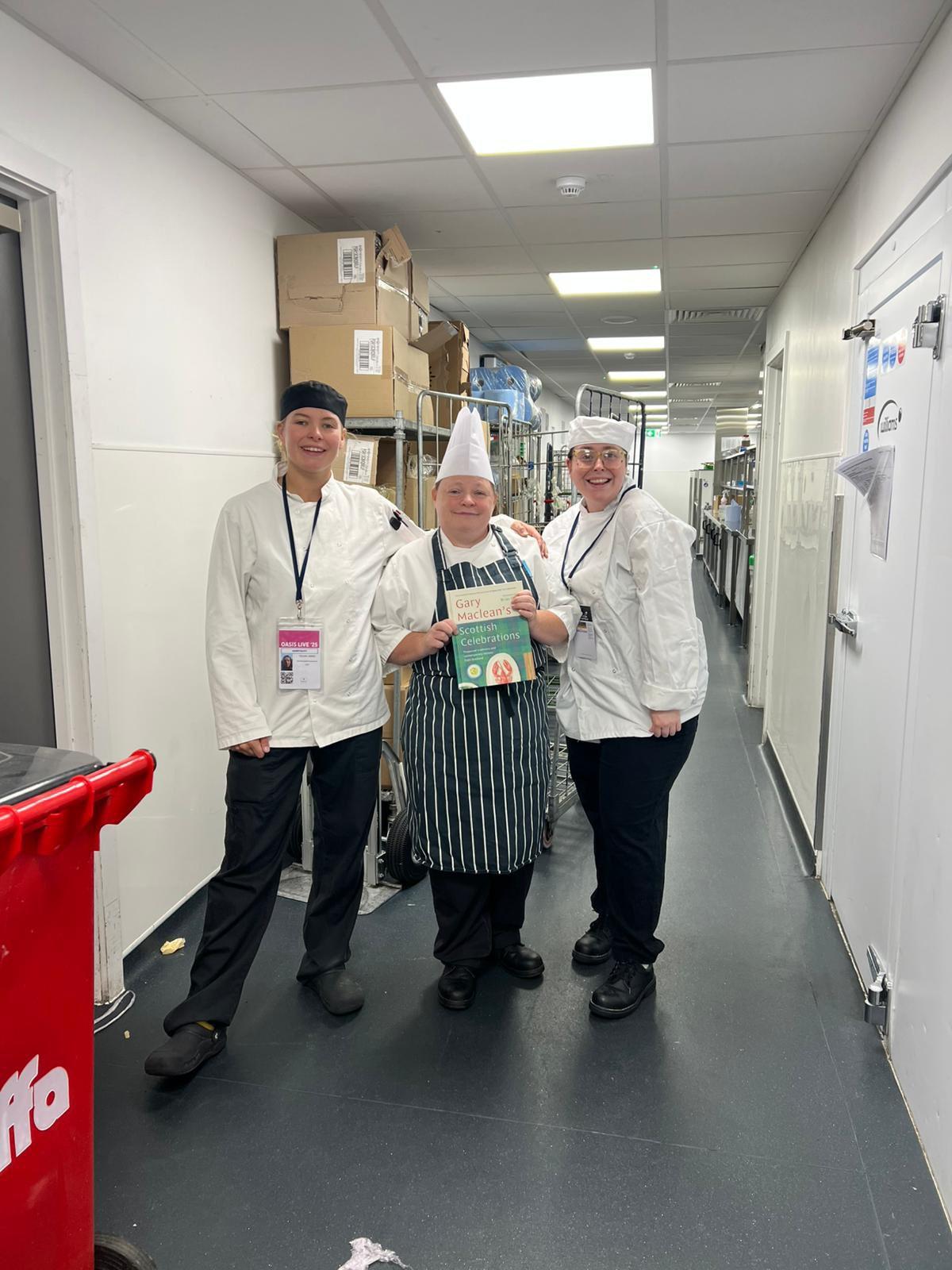 Three people in chef attire are standing in a storage corridor lined with shelves and boxes. The person in the middle is wearing a striped apron and holding a cookbook