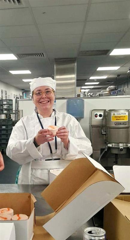A person wearing a white chef uniform and hat is standing in a commercial kitchen, holding a small dessert item. There are open cardboard boxes with similar desserts on a stainless steel counter in front of them. Industrial kitchen equipment and warning signage are visible in the background.