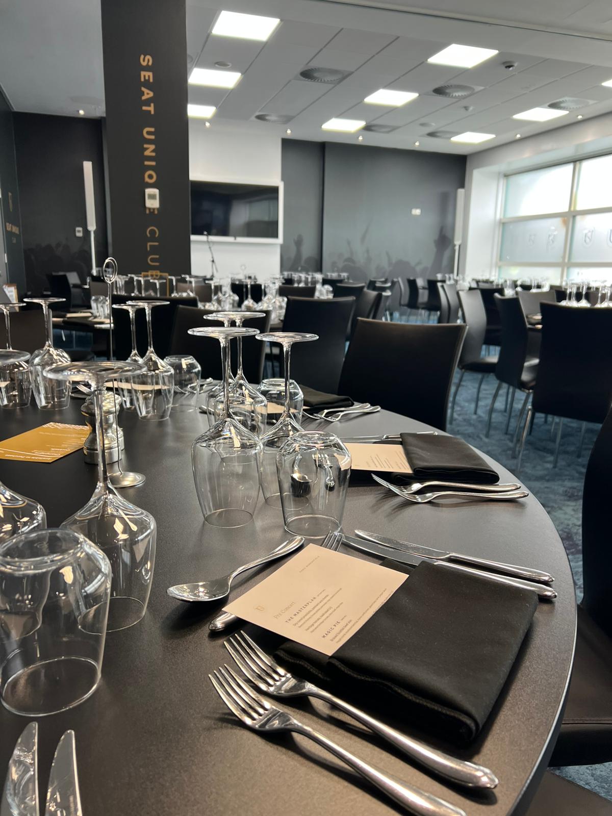 A modern dining area with round tables set for an event. Each table has neatly arranged wine glasses, cutlery, black napkins, and printed menus. The room features black chairs, large windows, and a wall with the words “SEAT UNION CLUB” in bold lettering.