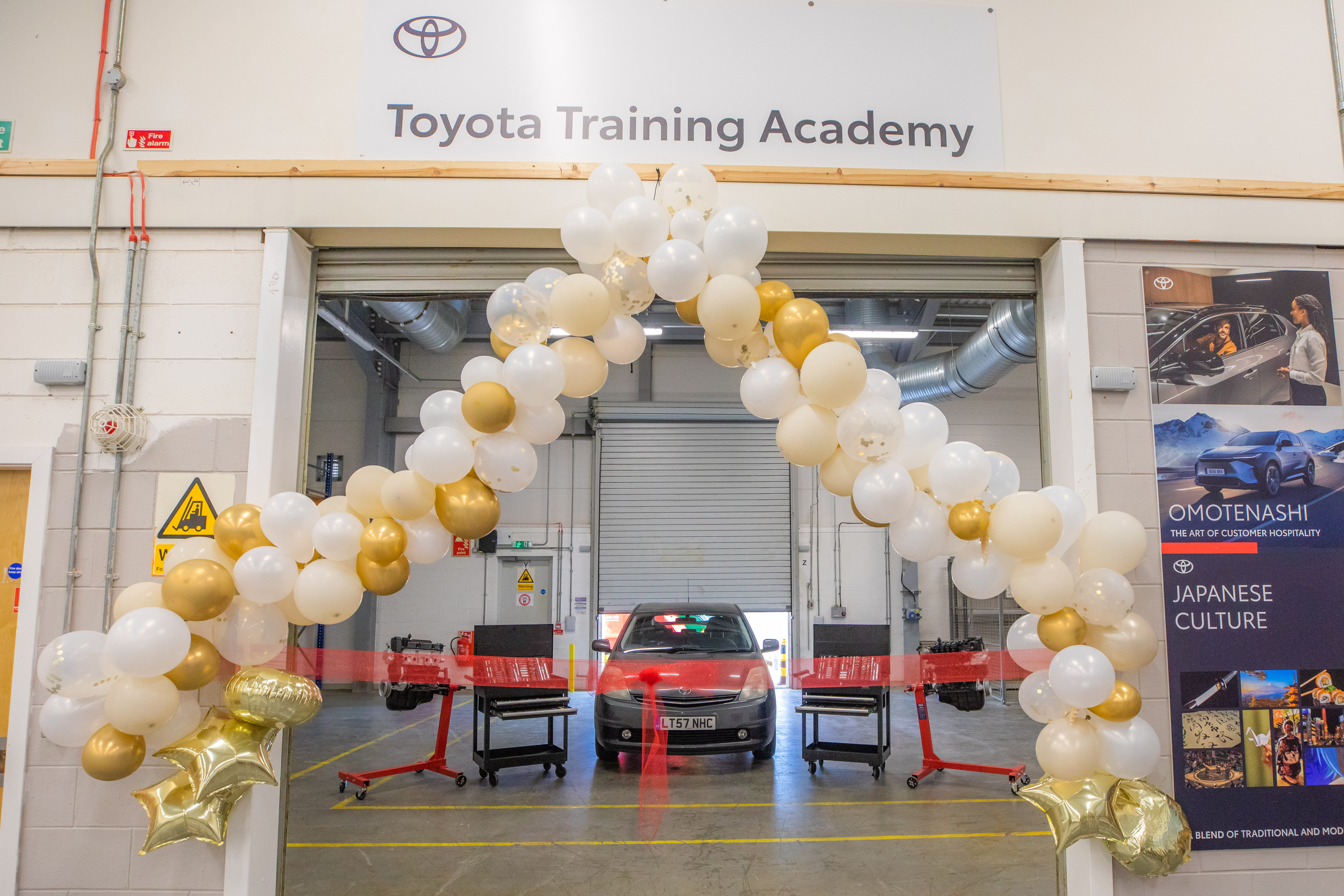Balloon arch in white and gold framing the entrance to the Toyota Training Academy, with a car visible inside and posters about Japanese culture on the wall.
