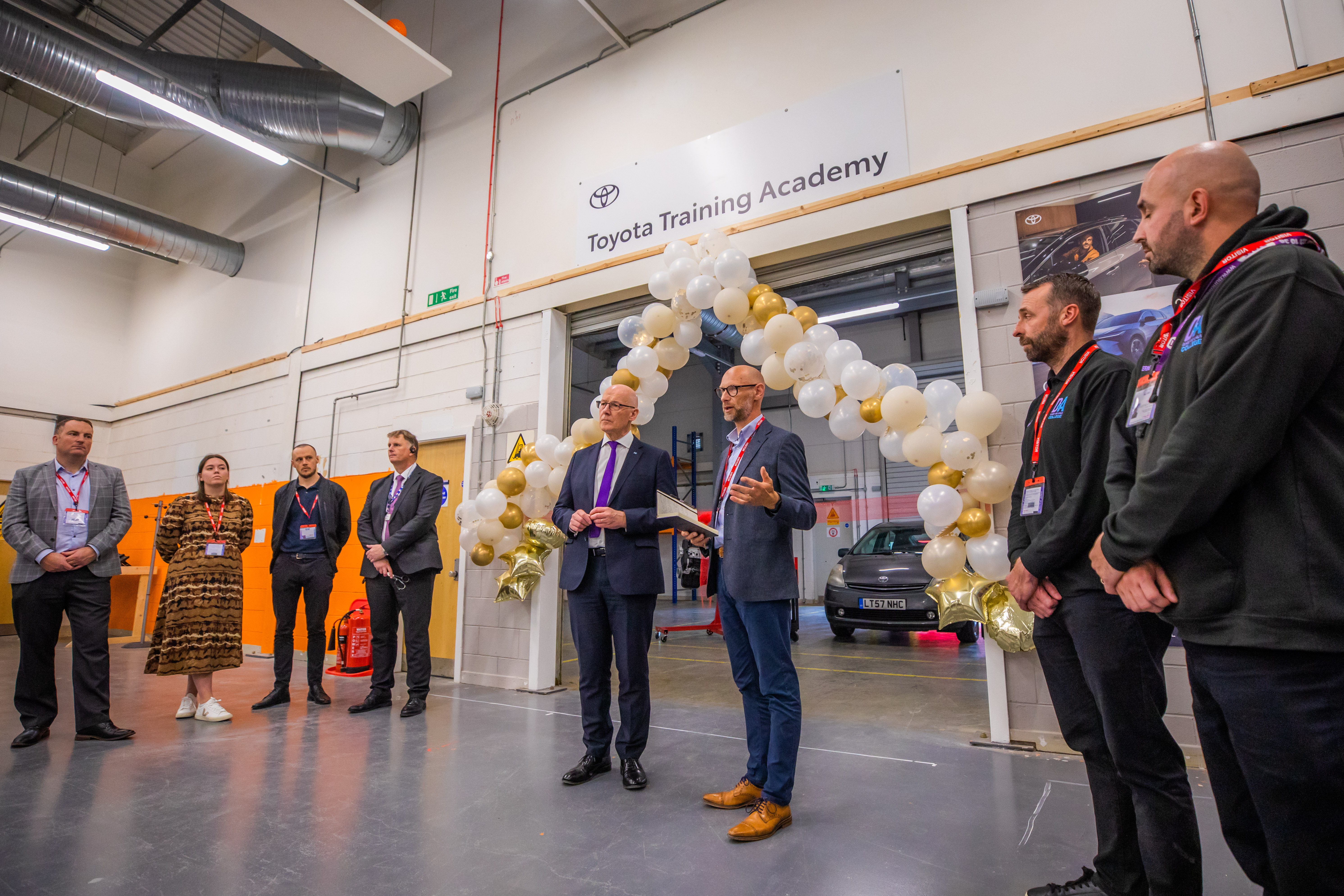 Group of people standing in a semi-circle inside a training facility, with a balloon arch and a sign reading "Toyota Training Academy" above an open doorway.)