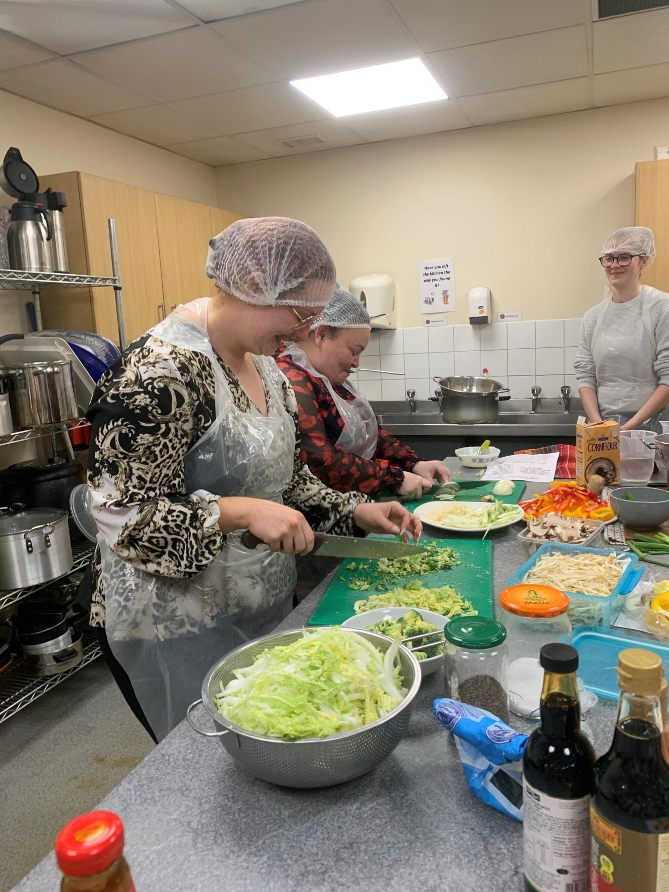 A kitchen setting with several people wearing hairnets and aprons, preparing food on a countertop. Various ingredients such as shredded lettuce, chopped vegetables, sauces, and condiments are spread across the counter. Large cooking pots are visible in the background.